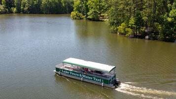 Un bateau vert et blanc, sur lequel on peut lire les mots « Excursions sur le lac et service de transport fluvial », se déplace sur un lac paisible, entouré d’une forêt dense.