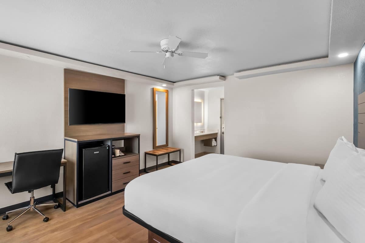 Habitación de hotel con cama, escritorio, silla, armario, espejo y ventilador de techo.