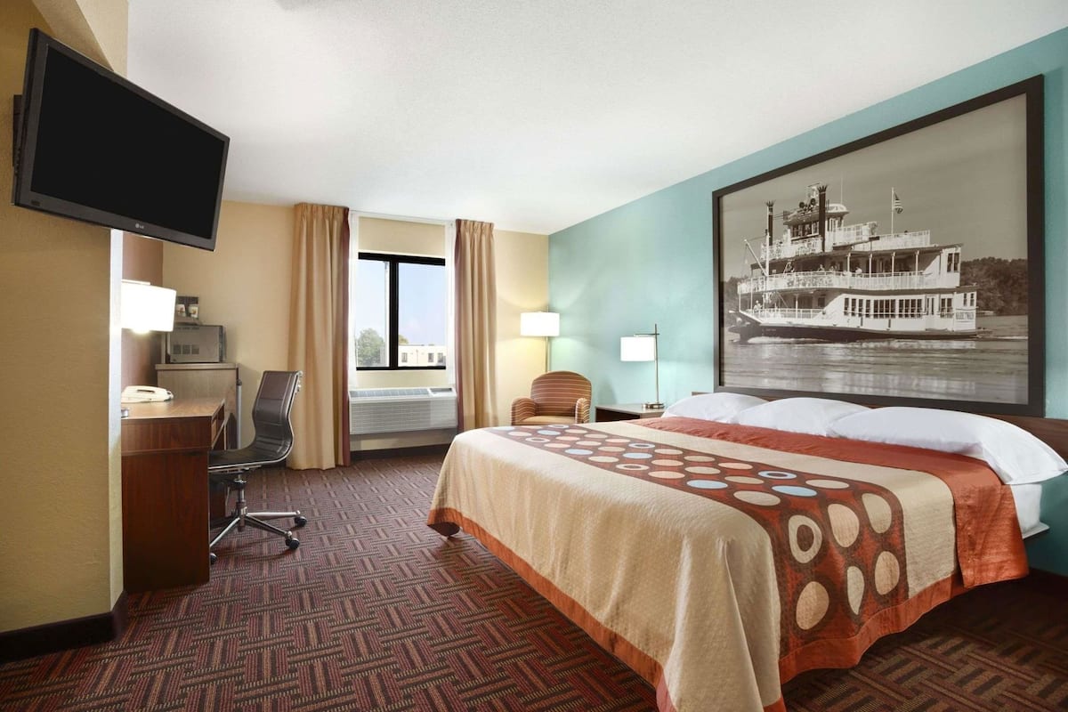 A hotel room with a bed, a desk with a computer, a television, a window with curtains, and a large picture of a boat on the wall.