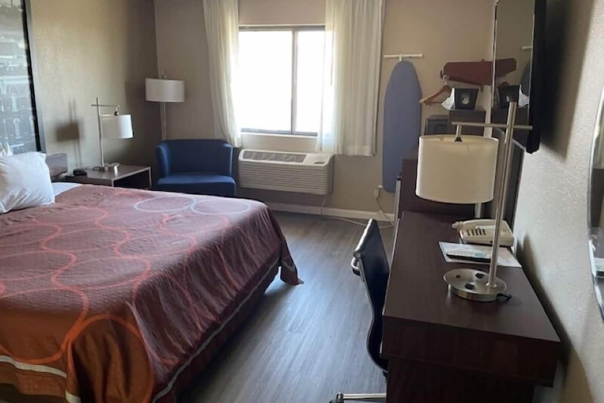 A hotel room with a large bed, a desk with a lamp, a chair, an ironing board, and a window with curtains.