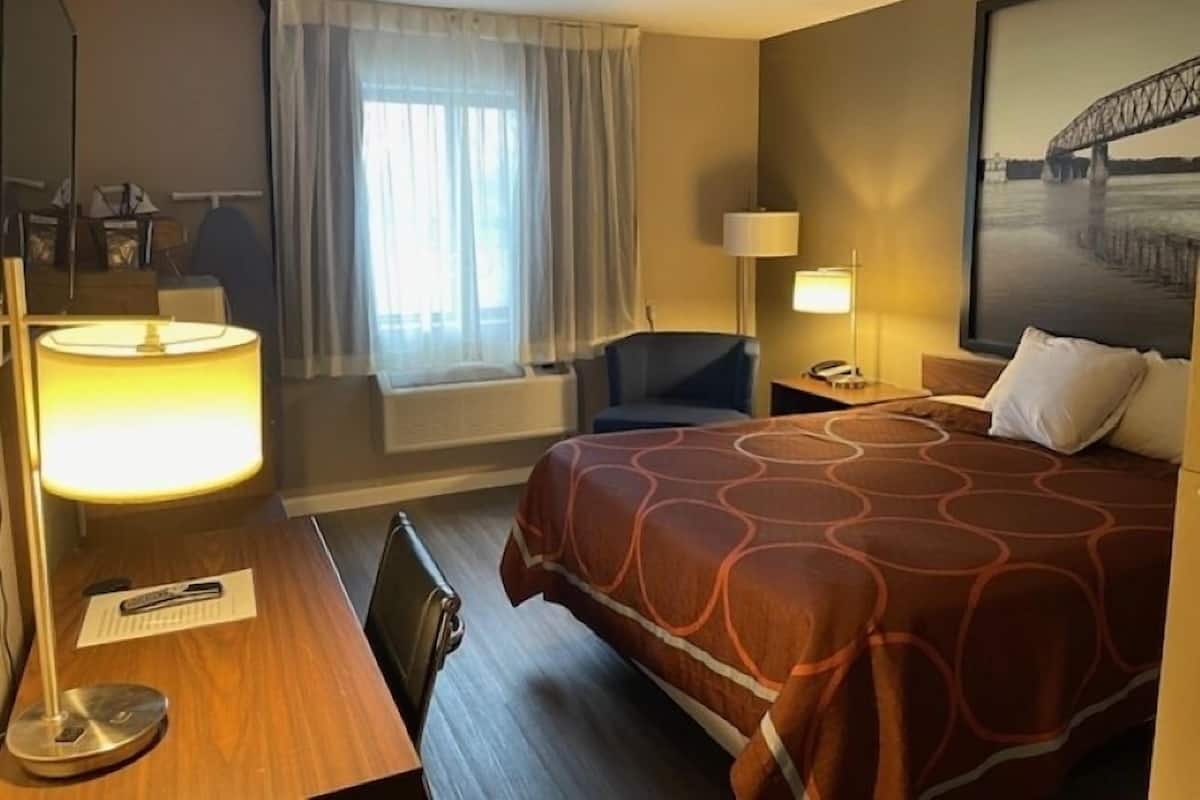 A hotel room with a large bed, a desk with a lamp, a chair, a window with curtains, and a painting on the wall.