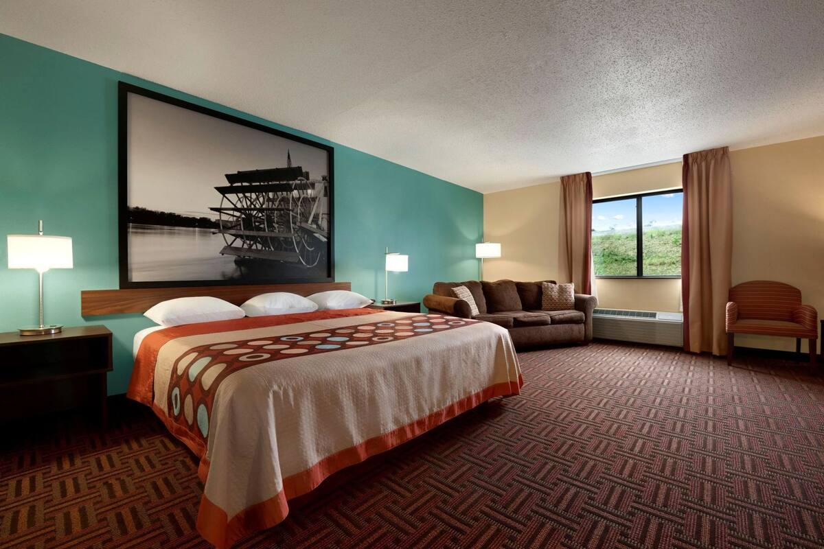 A hotel room with a large bed, a sofa, a chair, a window with a view, and a framed picture on the wall.