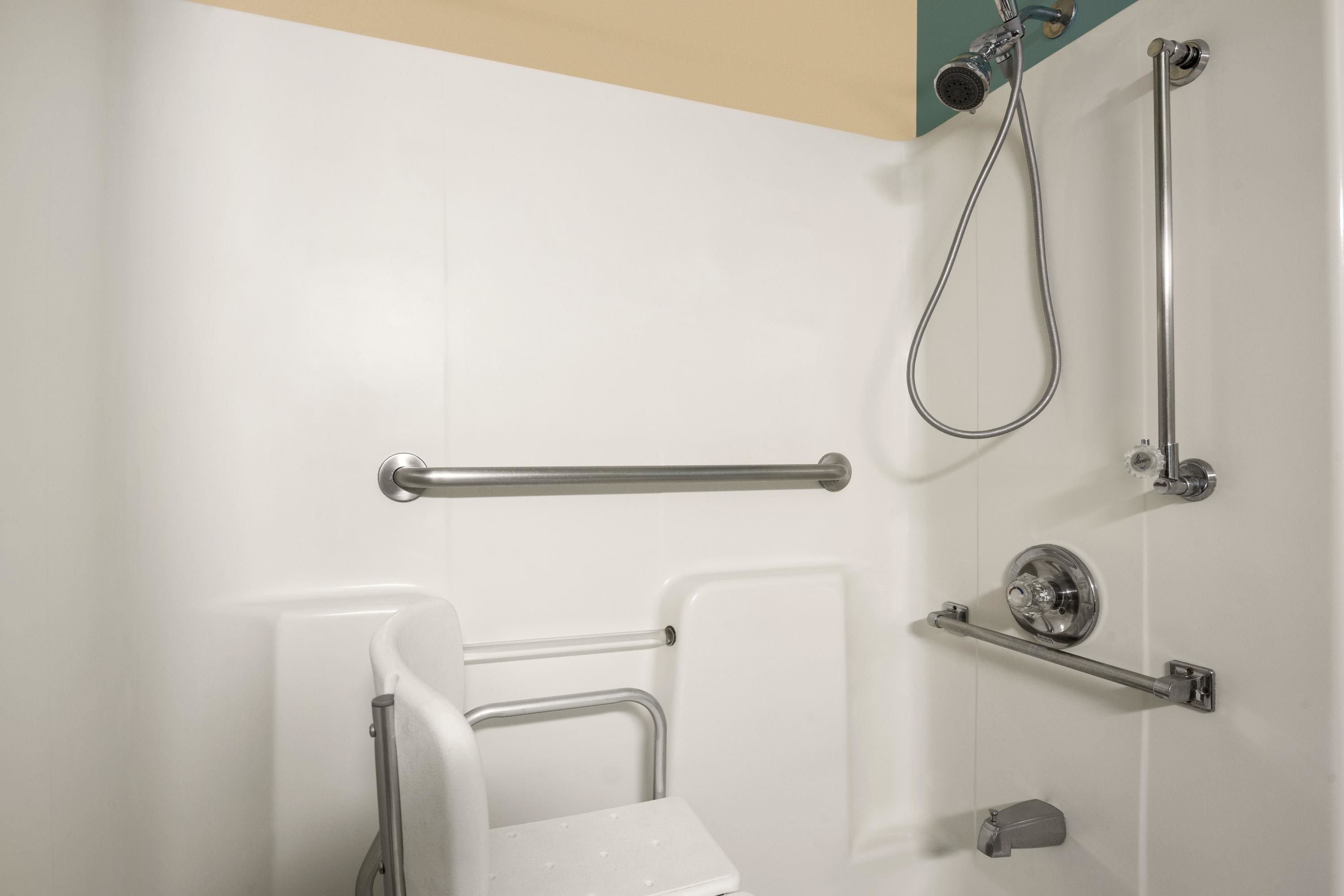 Un baño con pared de azulejos blancos, ducha con cabezal de ducha manual, barra de apoyo, inodoro con asiento de seguridad y jabonera.
