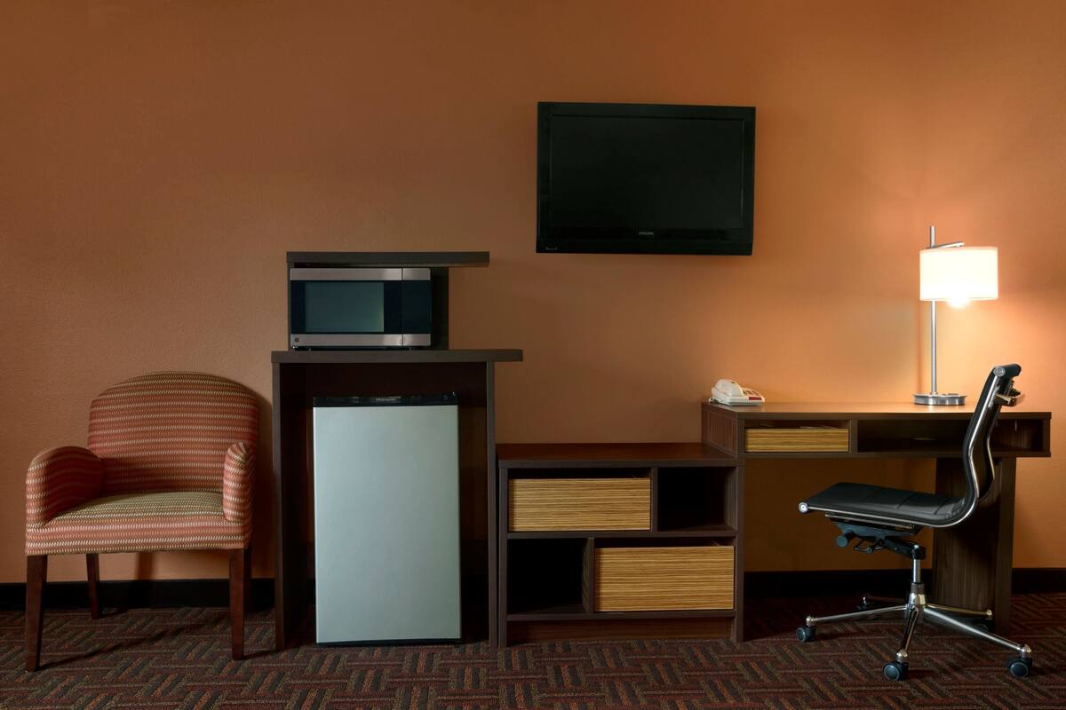 A hotel room with a desk, a chair, a television, a lamp, and a refrigerator.