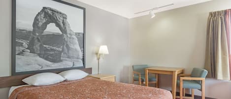 A hotel room with a large bed, a desk with two chairs, a lamp, and a framed photograph on the wall.