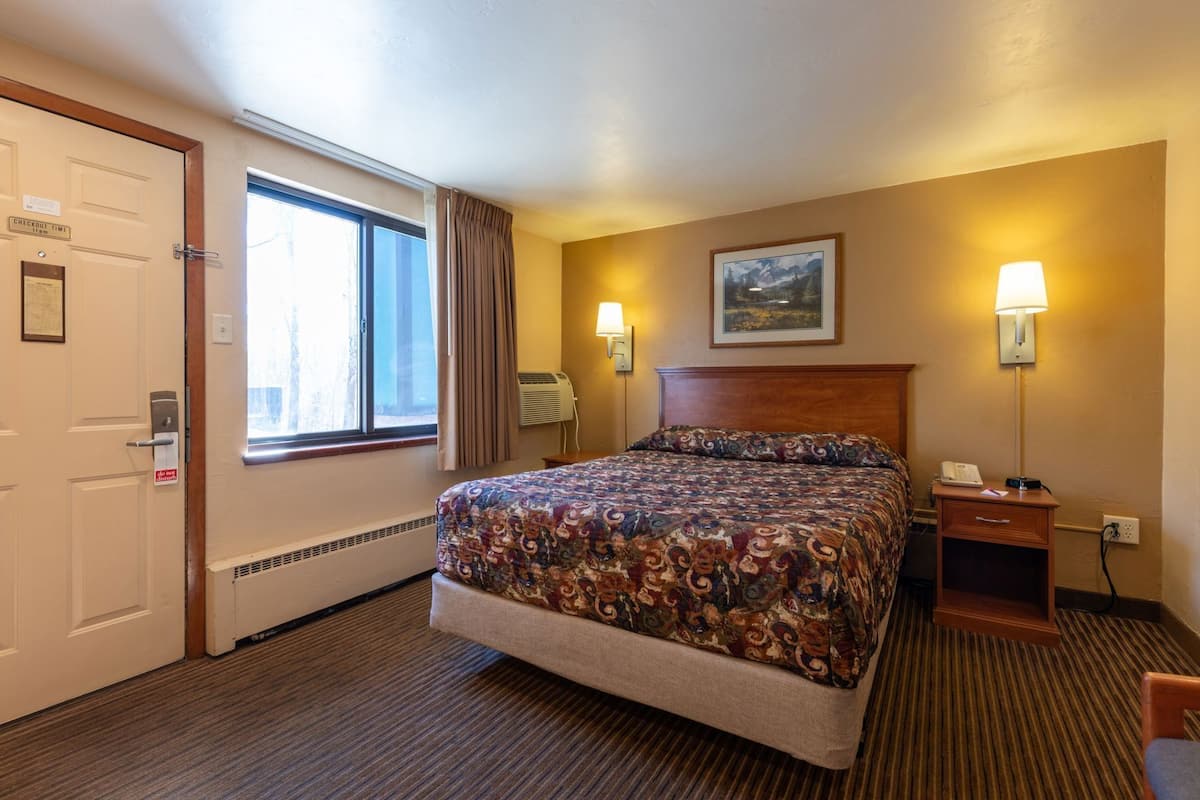 A hotel room with a bed, a nightstand, a lamp, a painting on the wall, and a window with curtains.