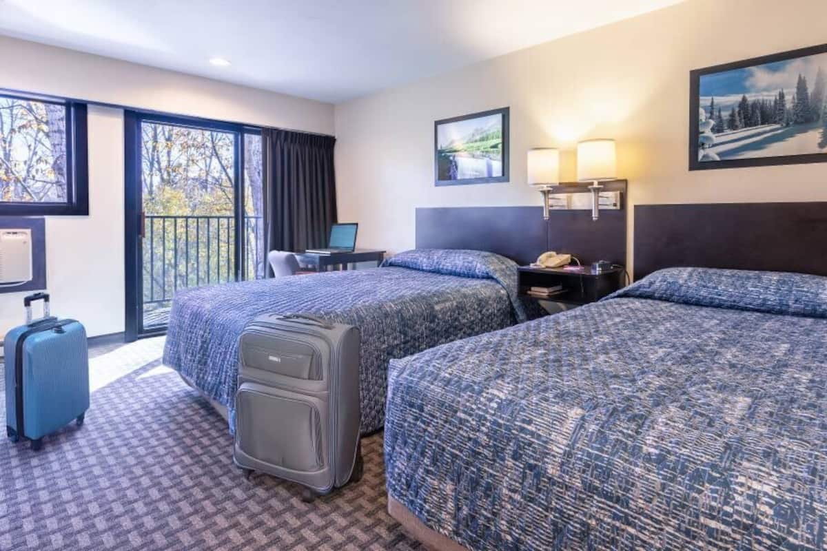 A hotel room with two beds, a desk, a chair, a suitcase, and a window with a view of trees.