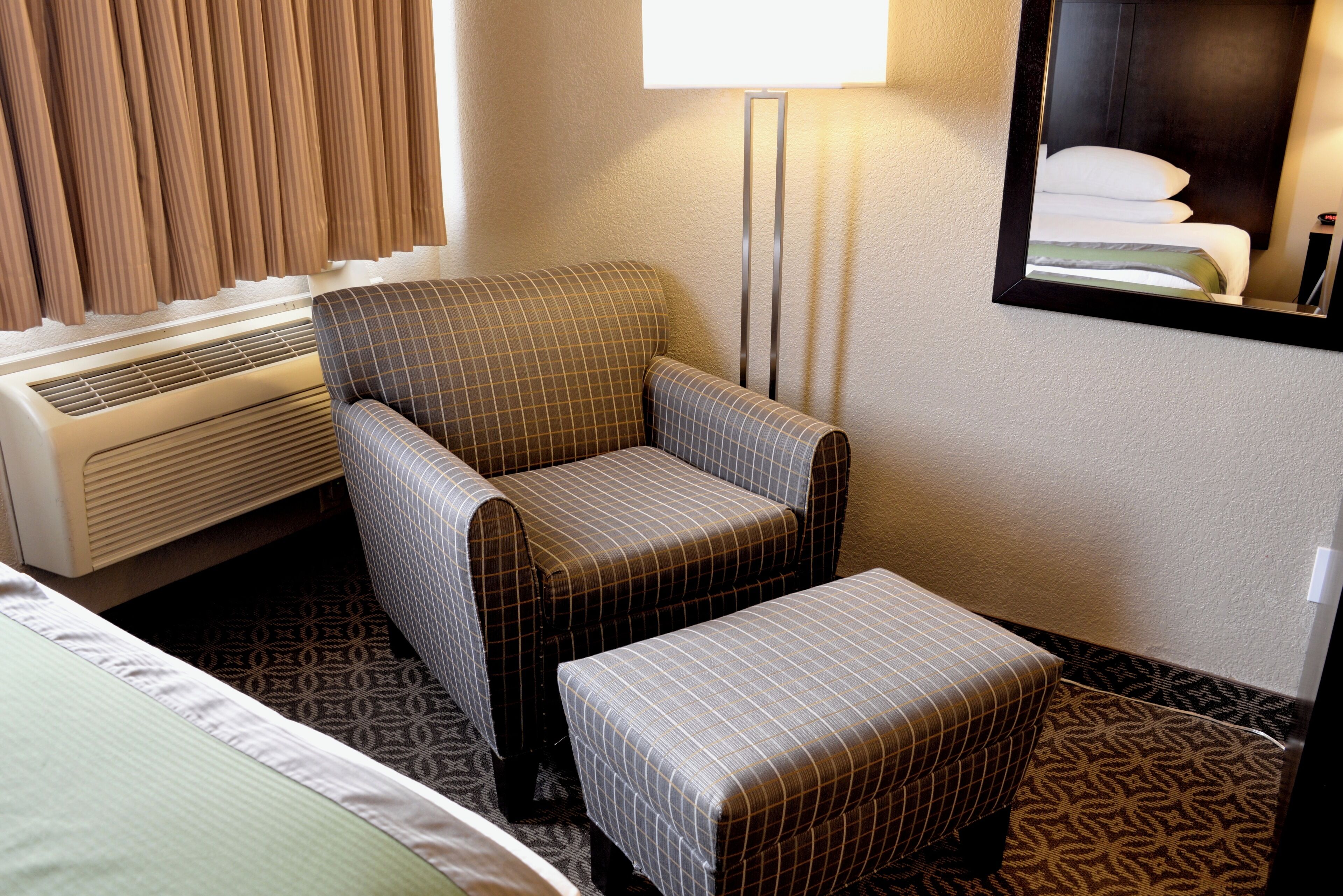 A hotel room with a bed, a chair, an ottoman, a standing lamp, and a window with curtains.