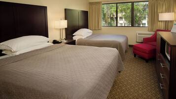 A hotel room with two beds, a red chair, a desk with a lamp, and a window with curtains.