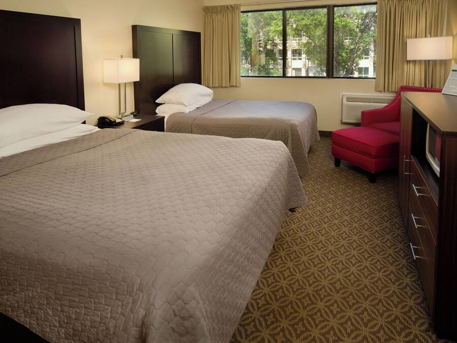 A hotel room with two beds, a red chair, a desk with a lamp, and a window with curtains.