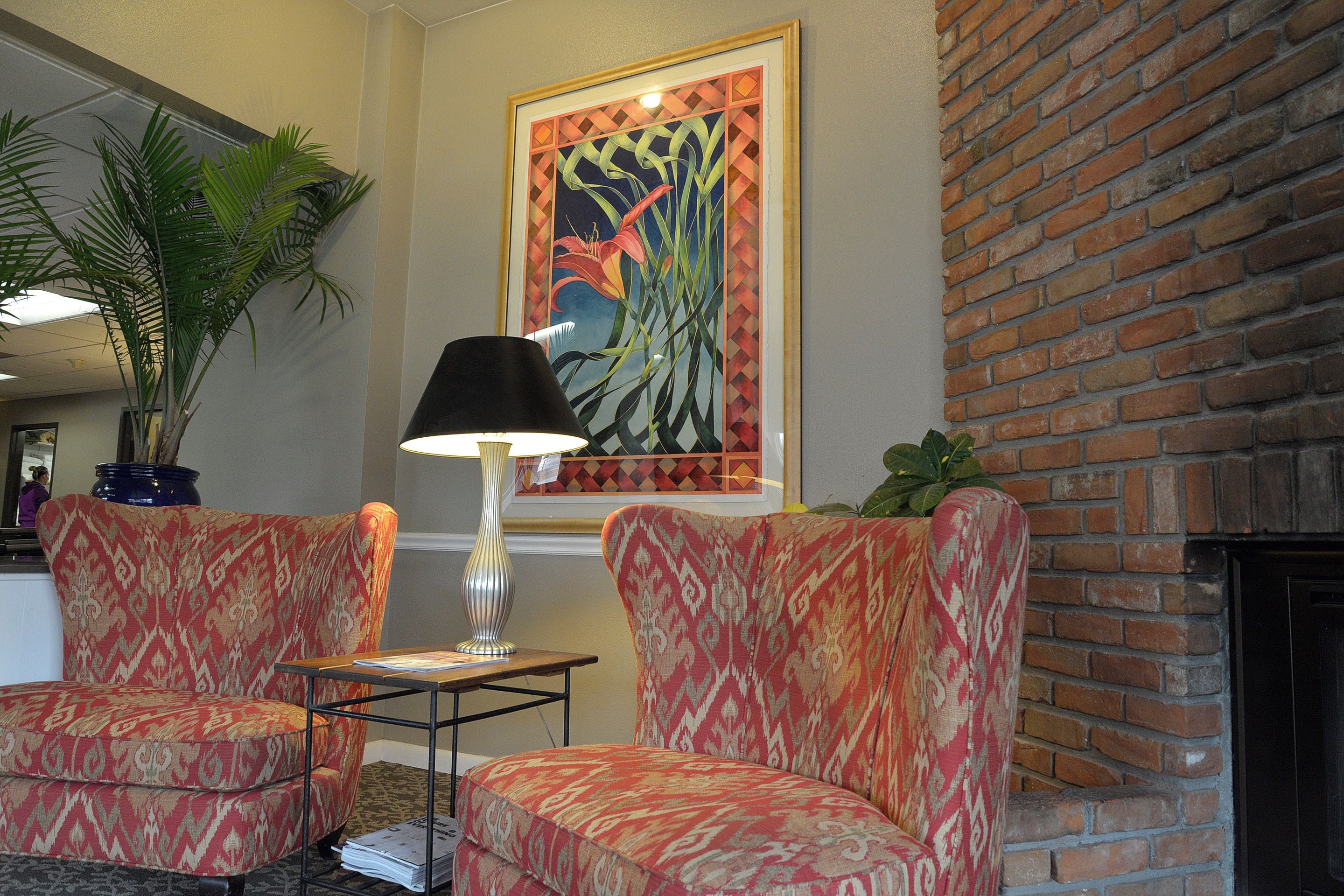 Un vestíbulo con dos sillones estampados, una mesa con lámpara, un cuadro grande enmarcado y una pared de ladrillo.