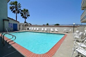 A rectangular swimming pool with a red brick border, surrounded by white lounge chairs and palm trees.