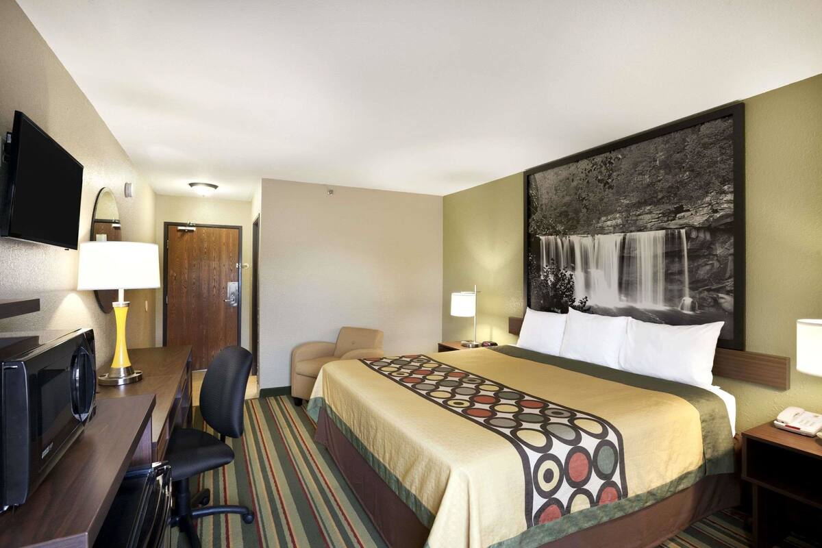 A hotel room with a bed, a desk with a television, a chair, a desk lamp, and a painting of a waterfall on the wall.