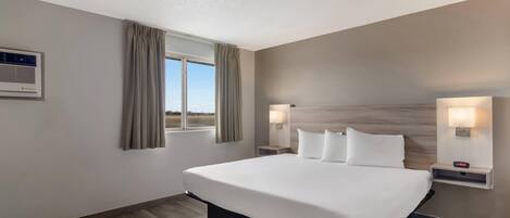 A hotel room with a large bed, two bedside tables with lamps, a window with curtains, and an air conditioning unit on the wall.