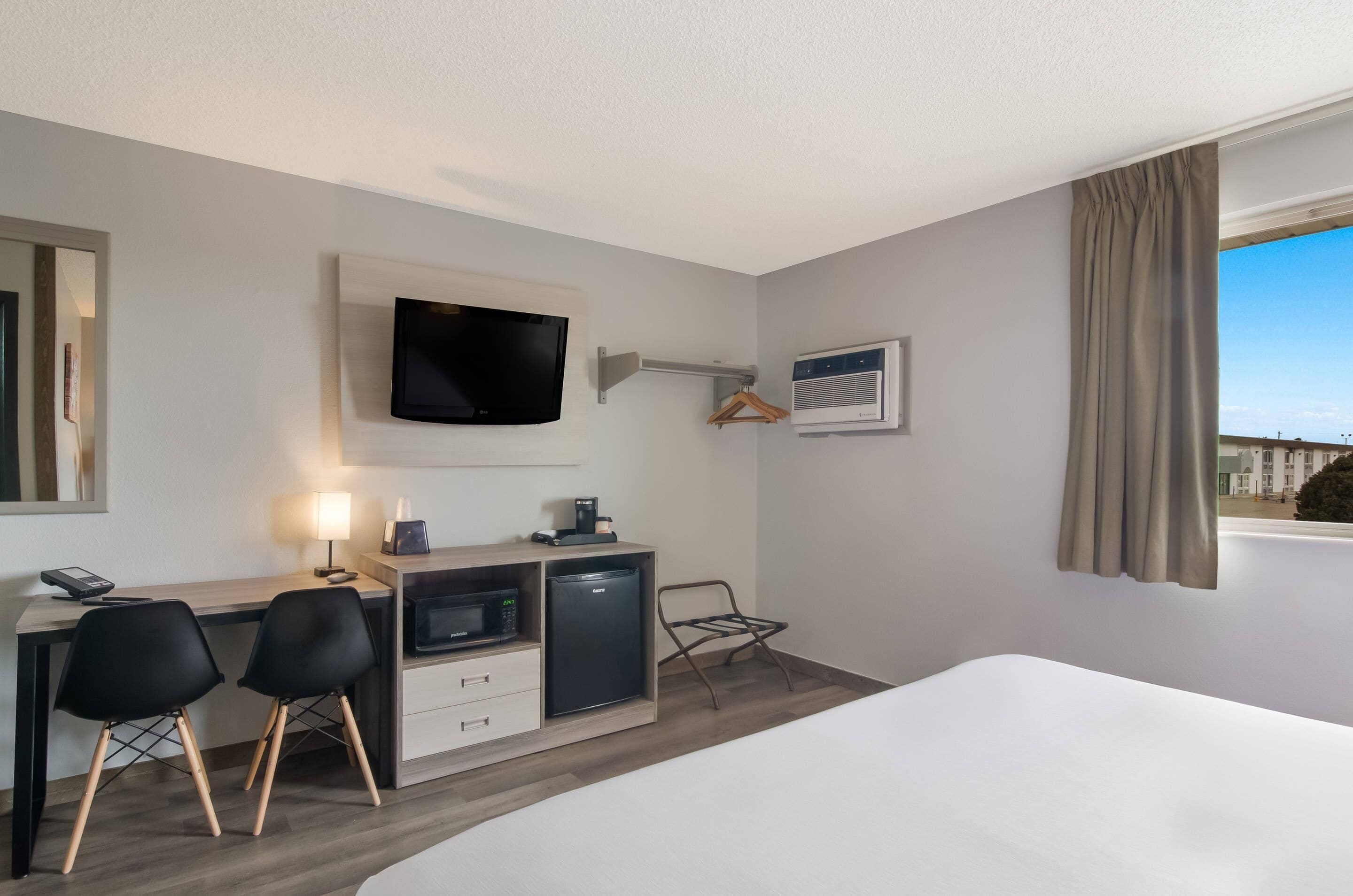 A hotel room with a bed, a desk with a computer, a microwave, and a refrigerator. There is a TV mounted on the wall, a window with curtains, and a chair.
