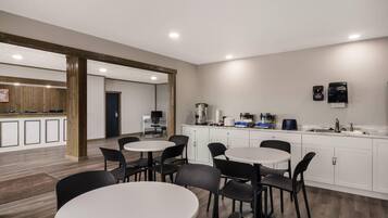 Une cuisine moderne avec des meubles blancs, un évier et une machine à café. On y trouve plusieurs tables et chaises rondes dans l’espace repas.