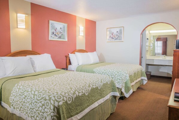 A hotel room with two beds, each with a green and white floral bedspread, a wooden headboard, and white pillows. There is a wooden nightstand with a lamp, a mirror, and a small table. The room has a red accent wall and a white ceiling. - Days Inn by Wyndham Bastrop (Bastrop)