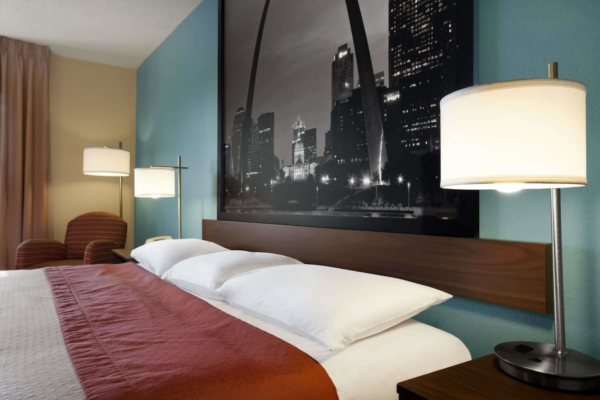 A hotel room with a bed, two bedside lamps, a cityscape painting, a wooden headboard, and a brown chair.