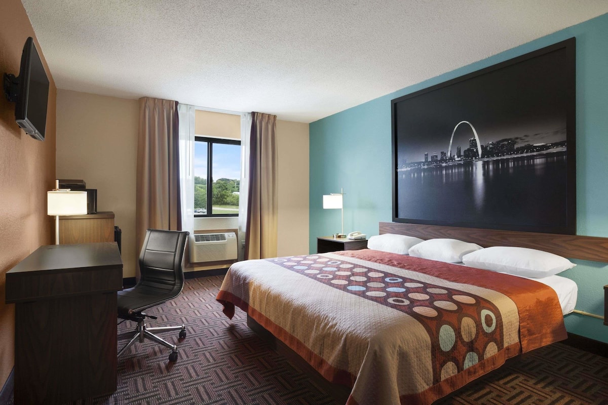 A hotel room with a bed, a desk, a chair, a TV, a window with curtains, and a cityscape painting.