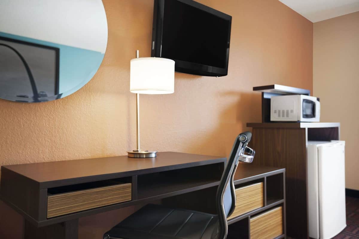 A hotel room with a desk, a television mounted on the wall, a lamp, a microwave, and a refrigerator.