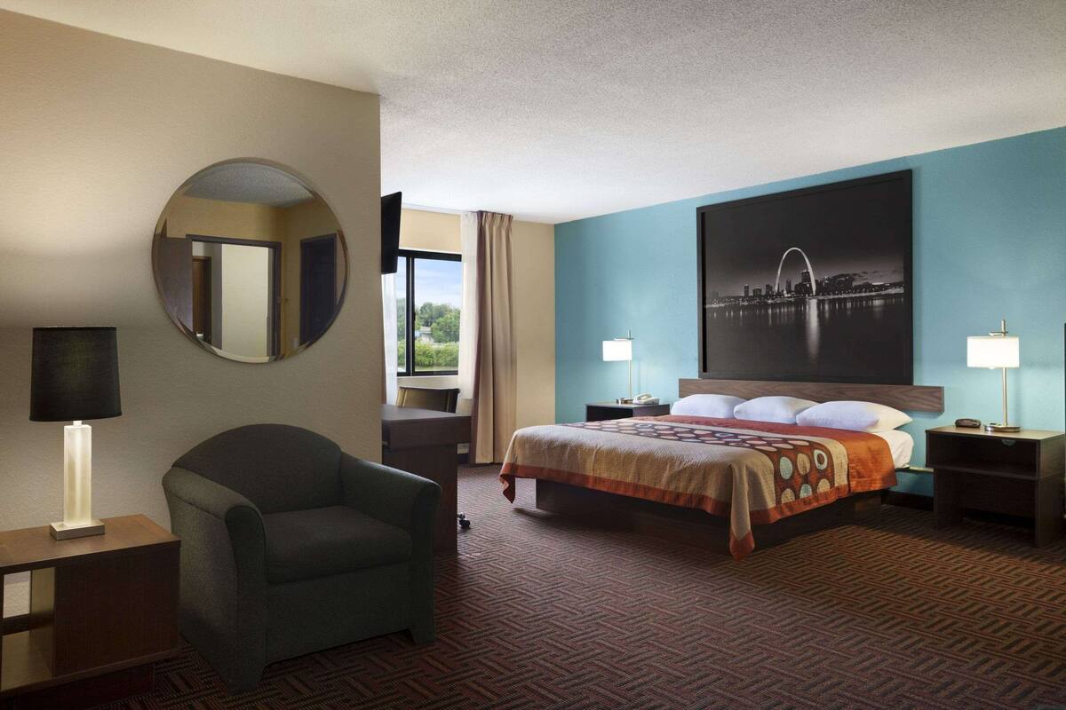 A hotel room with a large bed, a round mirror, a chair, a desk, and a nightstand with lamps.