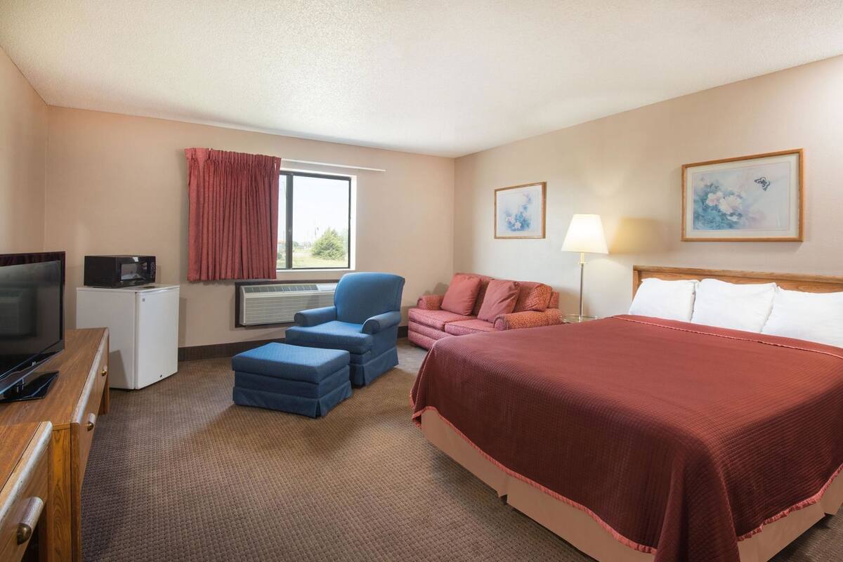 A hotel room with a bed, a desk with a TV, a chair, a couch, and a window with curtains.