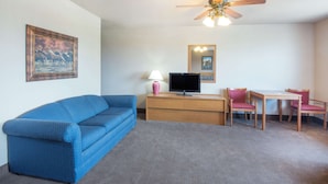 A room with a blue sofa, a wooden desk with a TV, a chair, a ceiling fan, and a framed picture on the wall. - Super 8 by Wyndham Abilene KS (Abilene)