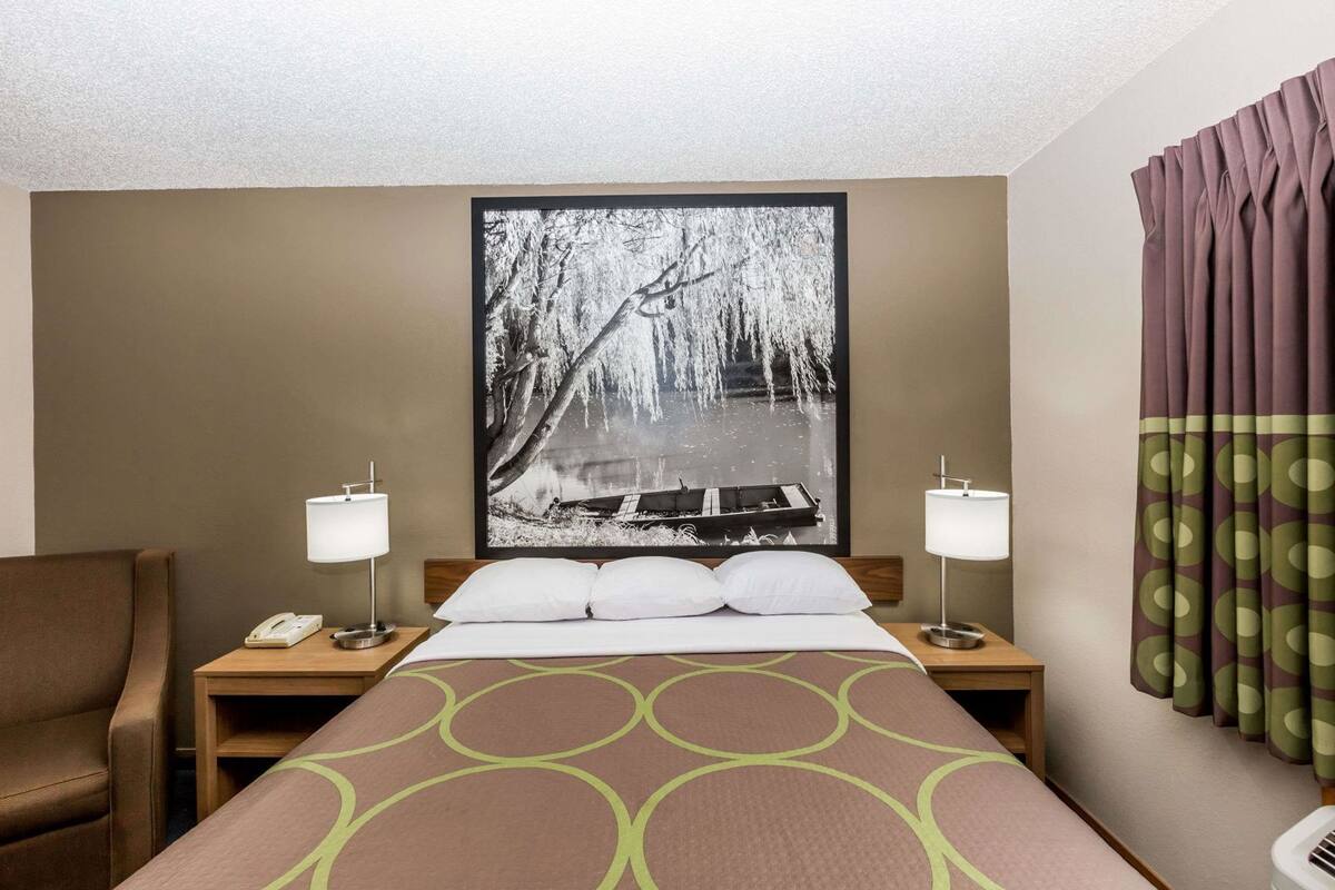 Een hotelkamer met een bed, twee nachtkastjes, een telefoon, een lamp, een groot fotokader en een gordijn.