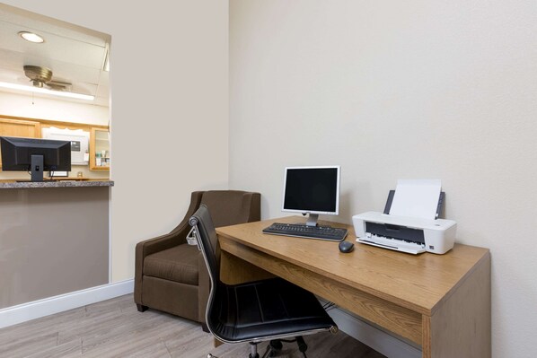 Habitación de hotel con un escritorio de madera, una computadora, una impresora, un sillón marrón y una pequeña zona de kitchenette.