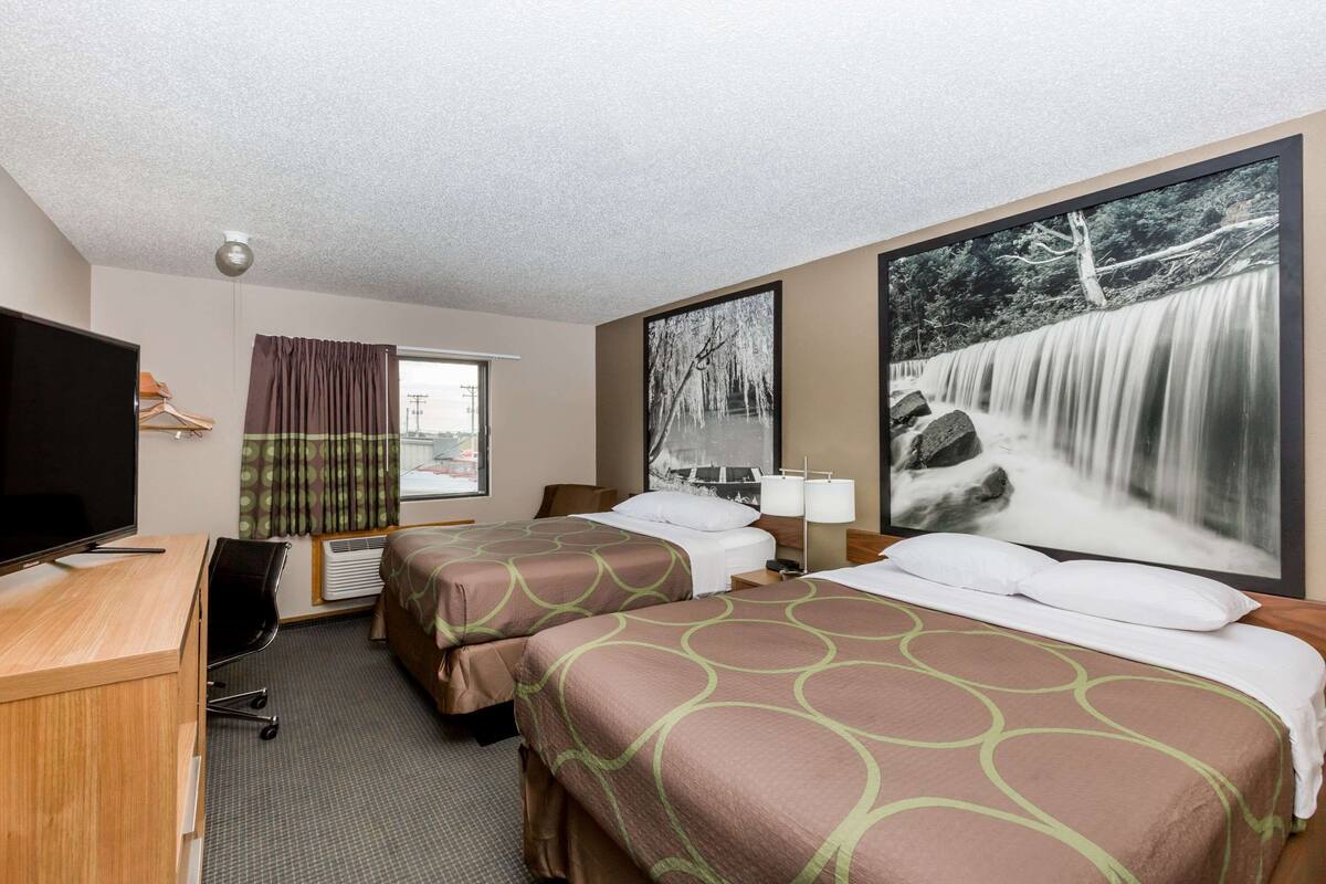 Een hotelkamer met twee bedden, een bureau met een televisie, een raam met gordijnen, en een groot schilderij van een waterval aan de muur.