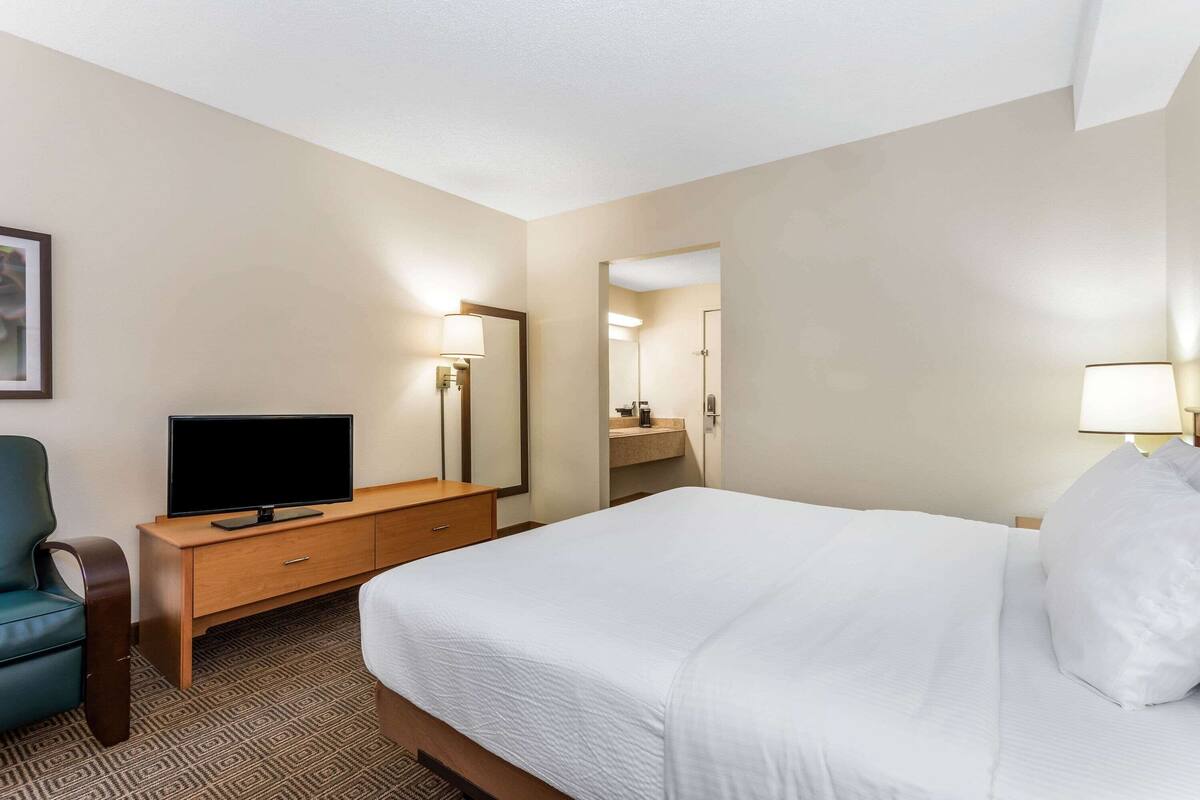 A hotel room with a bed, a TV on a wooden dresser, a chair, and a bathroom visible through an open door.