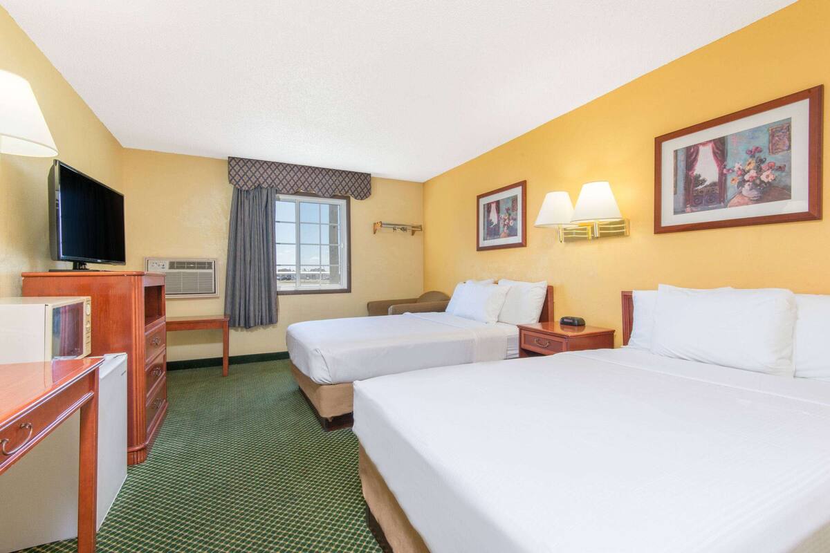 A hotel room with two beds, a desk with a television, a microwave, and a window with curtains.