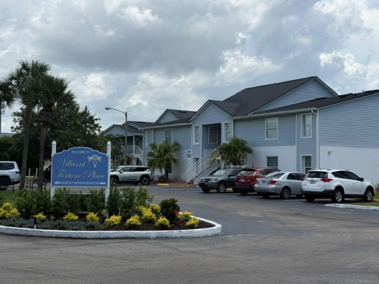 Um hotel com placa de registro de hóspedes, vários carros estacionados e uma área ajardinada com plantas pequenas.