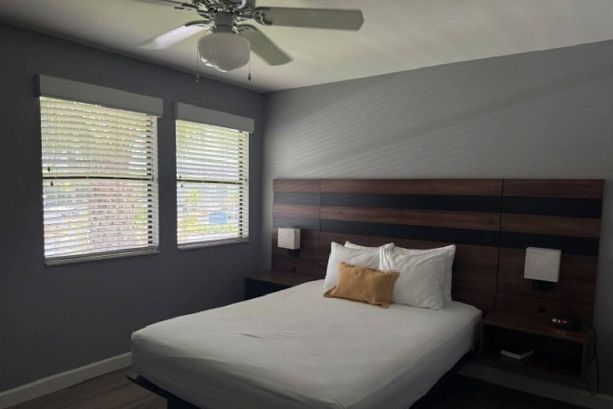 A bedroom with a bed, two windows with blinds, a ceiling fan, and a wooden headboard.