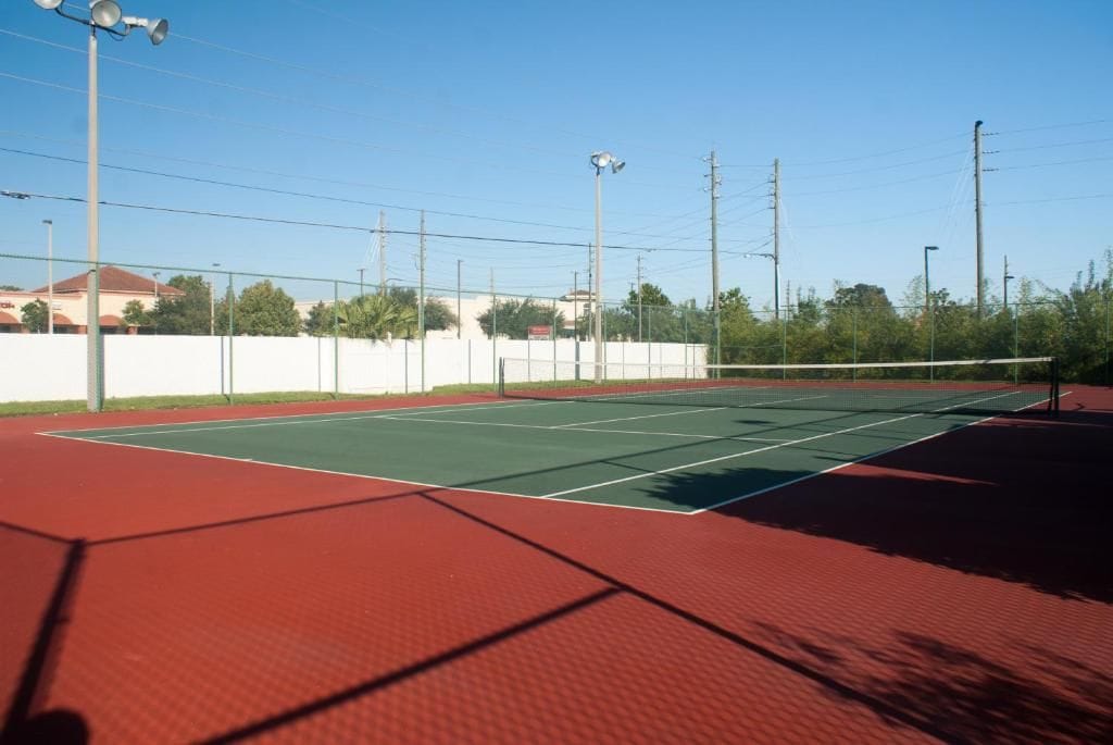 An outdoor tennis court with a red surface, surrounded by a white wall and trees.