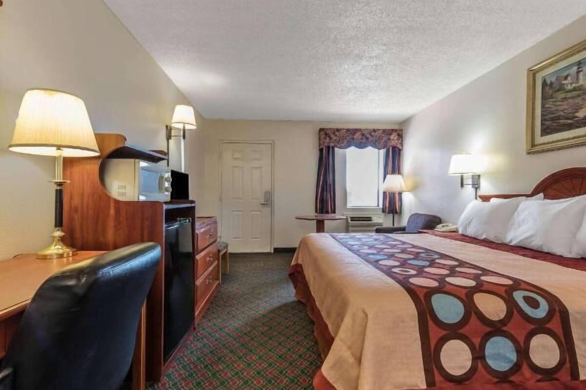 A hotel room with a bed, desk, chair, lamps, and a painting on the wall.