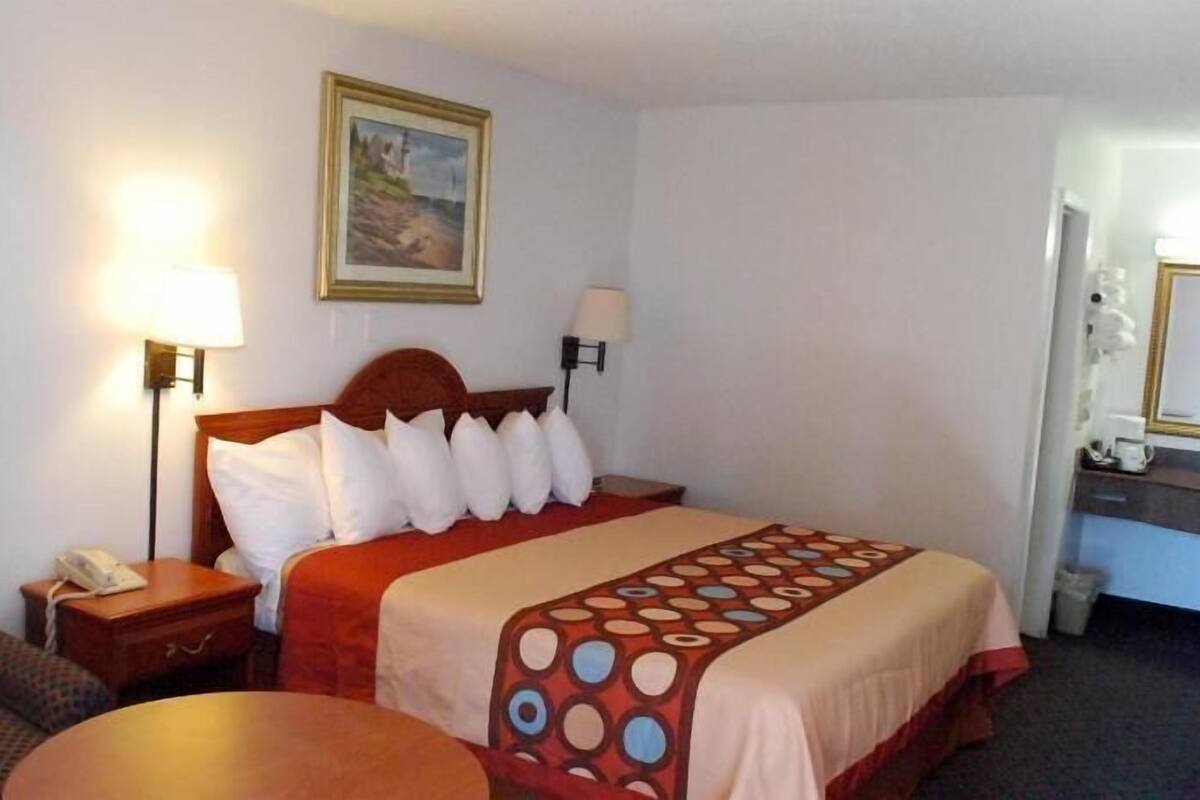 A hotel room with a bed, a chair, a small table, a nightstand, a lamp, a painting, and a mirror.