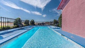 Una piscina rectangular amb un cantó blau de rajola, envoltada per una tanca de metall negre, i adjacent a un edifici amb una paret rosa.