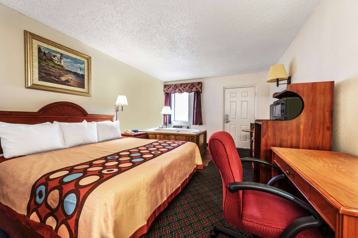 A hotel room with a bed, desk, chair, lamp, and a painting on the wall.