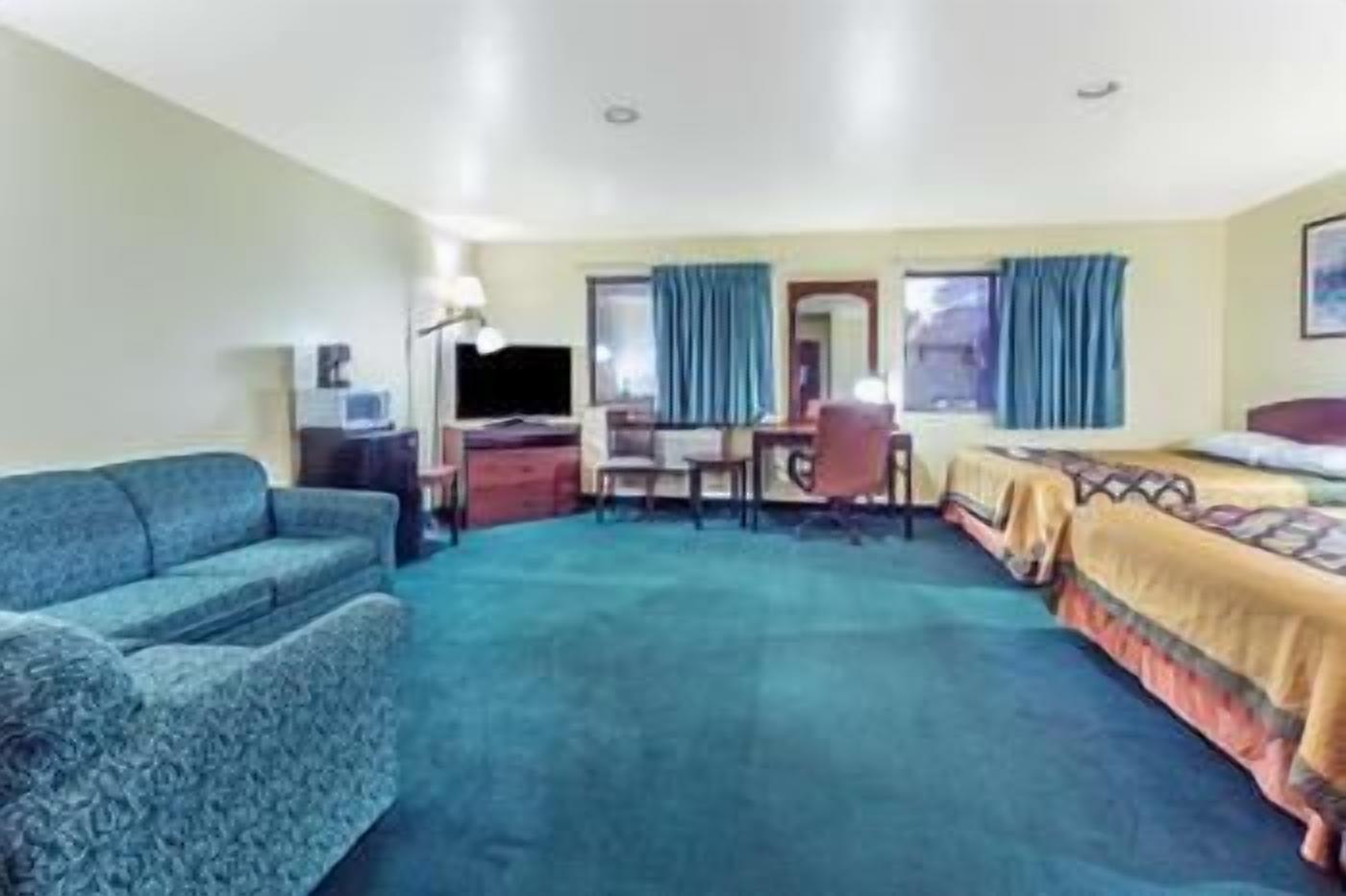 A hotel room with a blue carpet, a bed with a patterned bedspread, a desk with a chair, and a window with blue curtains.