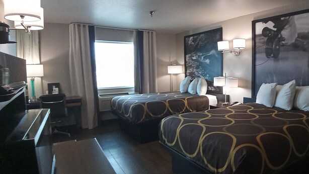 A hotel room with two beds, a desk, a chair, a window with curtains, and wall-mounted lamps.