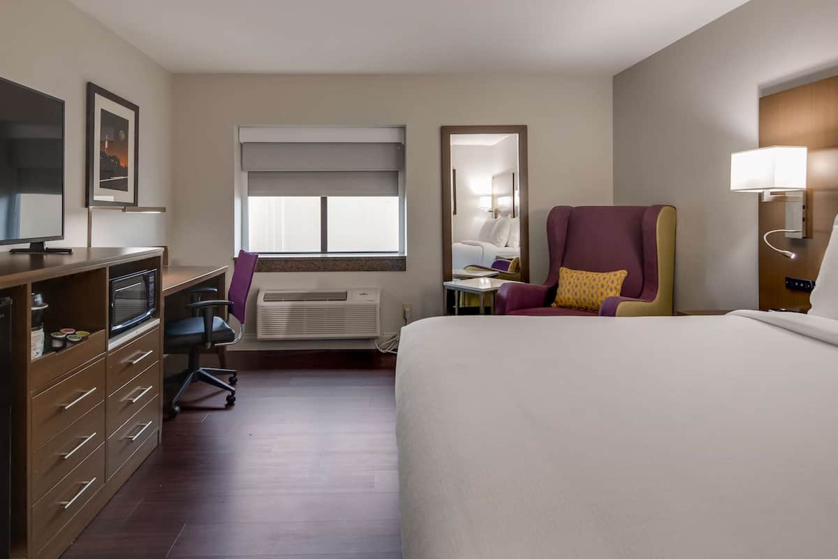 Ein Hotelzimmer mit einem Bett, einem Schreibtisch mit Computer, einem Sessel, einem Fernseher, einem Fenster mit Jalousien und einem Spiegel.