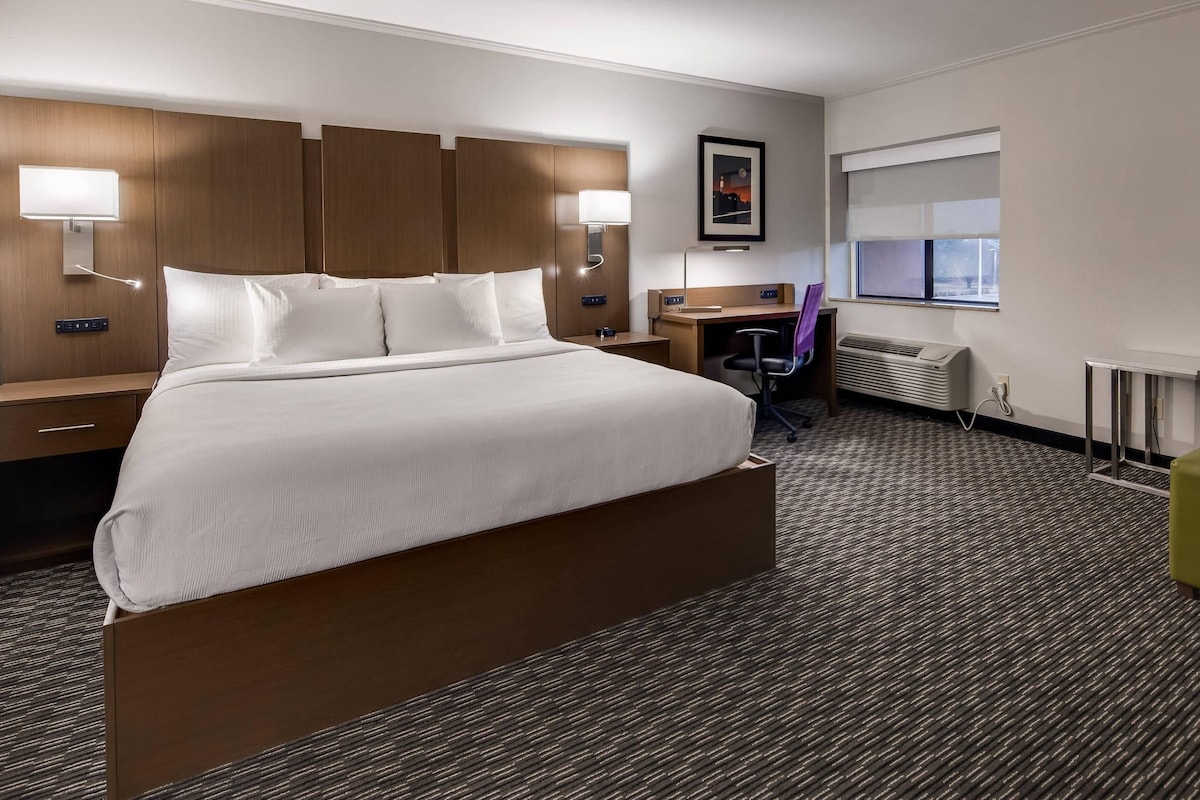 A hotel room with a large bed, a desk, a chair, and a small green ottoman.