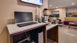 Una habitación de hotel moderna con un escritorio que tiene una computadora portátil, teclado y ratón. Hay tazas apiladas, una cafetera y un bote de basura. Un sofá y una ventana con vista a la calle son visibles al fondo.
