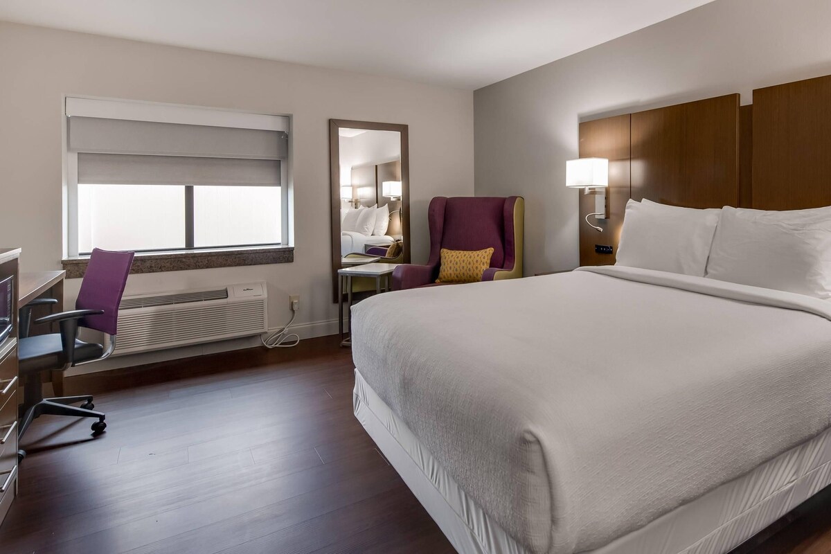 A hotel room with a large bed, a desk, a chair, a window with blinds, a heater, and a nightstand with a lamp.