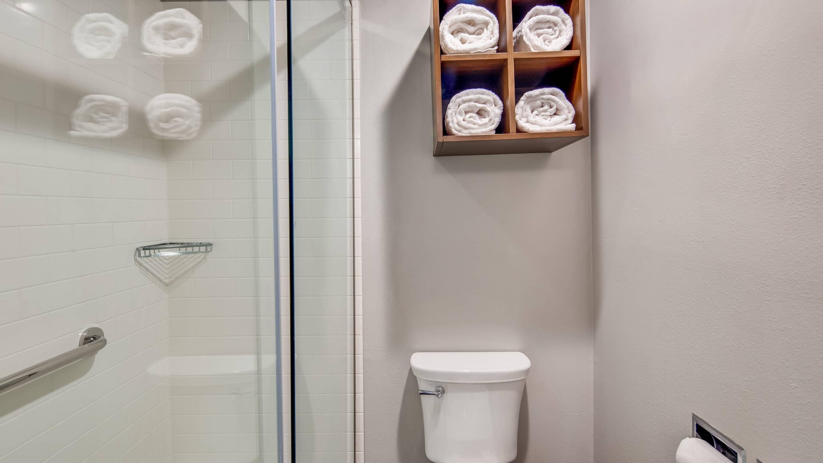 A bathroom with a glass shower enclosure, a toilet, and a shelf with rolled towels.