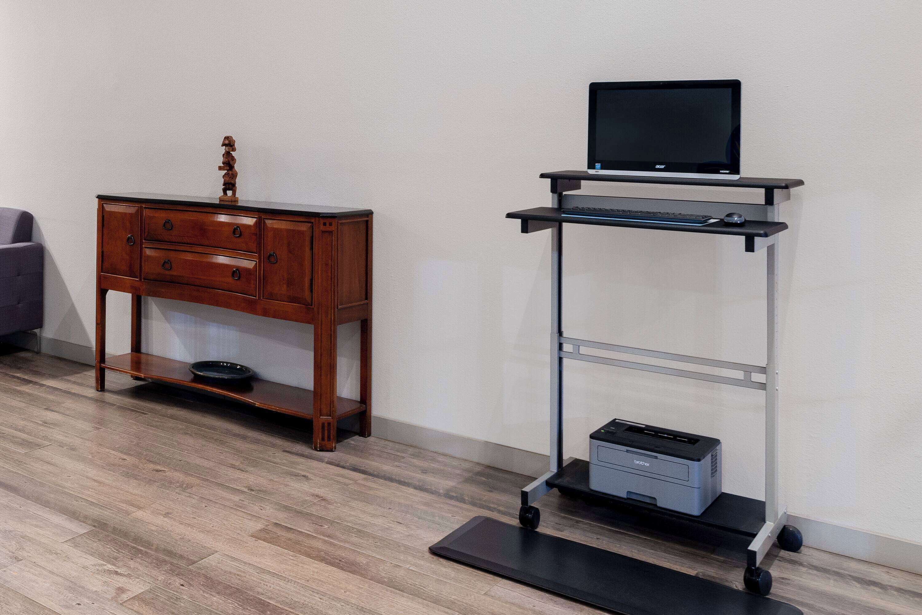 Une commode en bois avec des tiroirs et une petite étagère, un bureau noir avec un écran d’ordinateur et un clavier, et une imprimante posée sur un chariot.