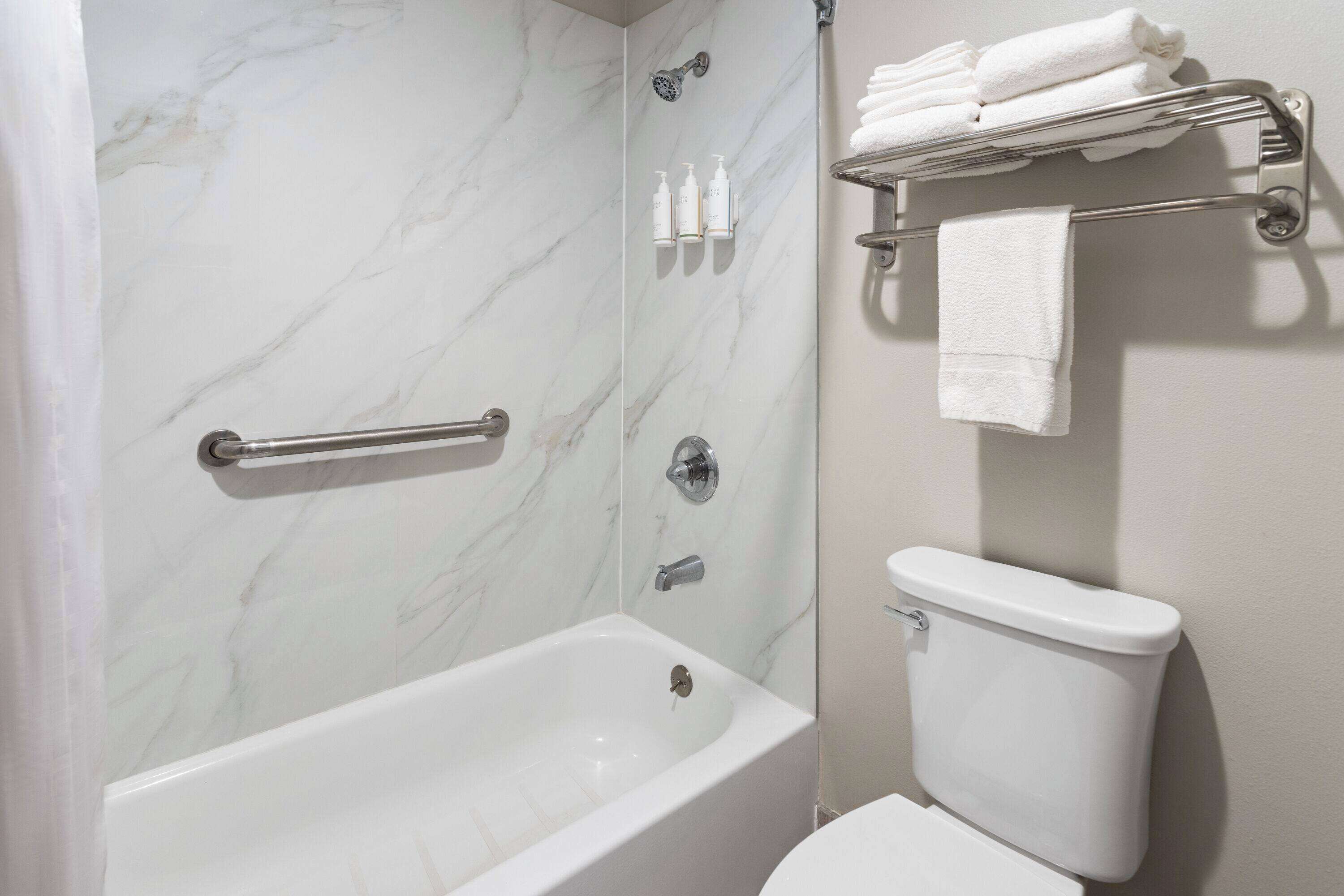 A bathroom with a marble shower wall, a bathtub, a toilet, and a towel rack with folded towels.