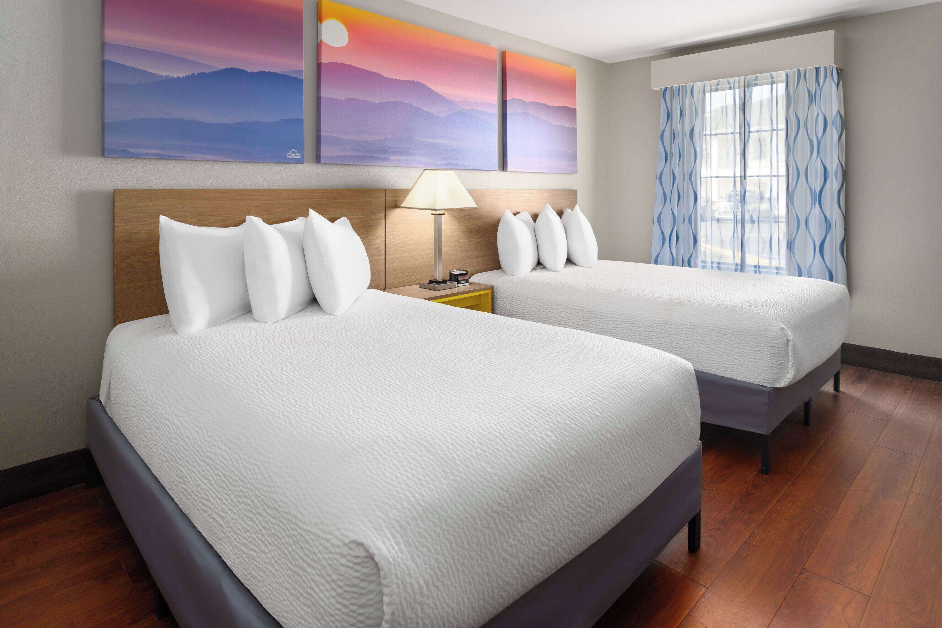 A hotel room with two beds, a nightstand with a lamp, and a window with patterned curtains.