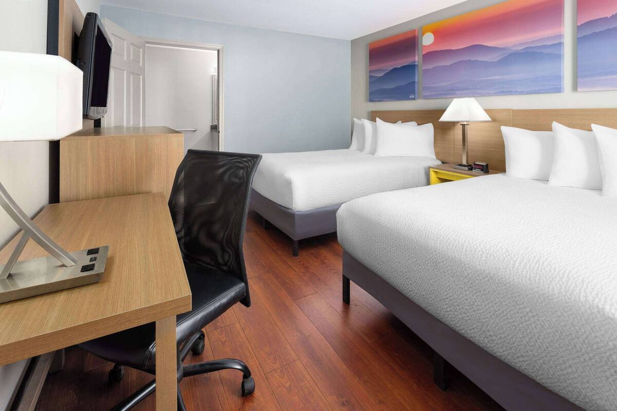 Quarto de hotel com duas camas, uma escrivaninha, uma cadeira, uma televisão e um quadro de pôr do sol.